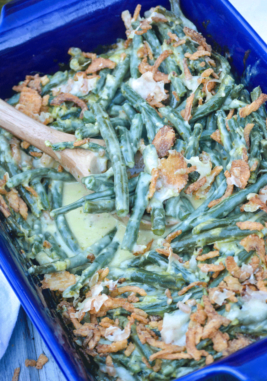 a wooden spoon lifting a scoop of basil pesto covered green beans out of a blue casserole dish