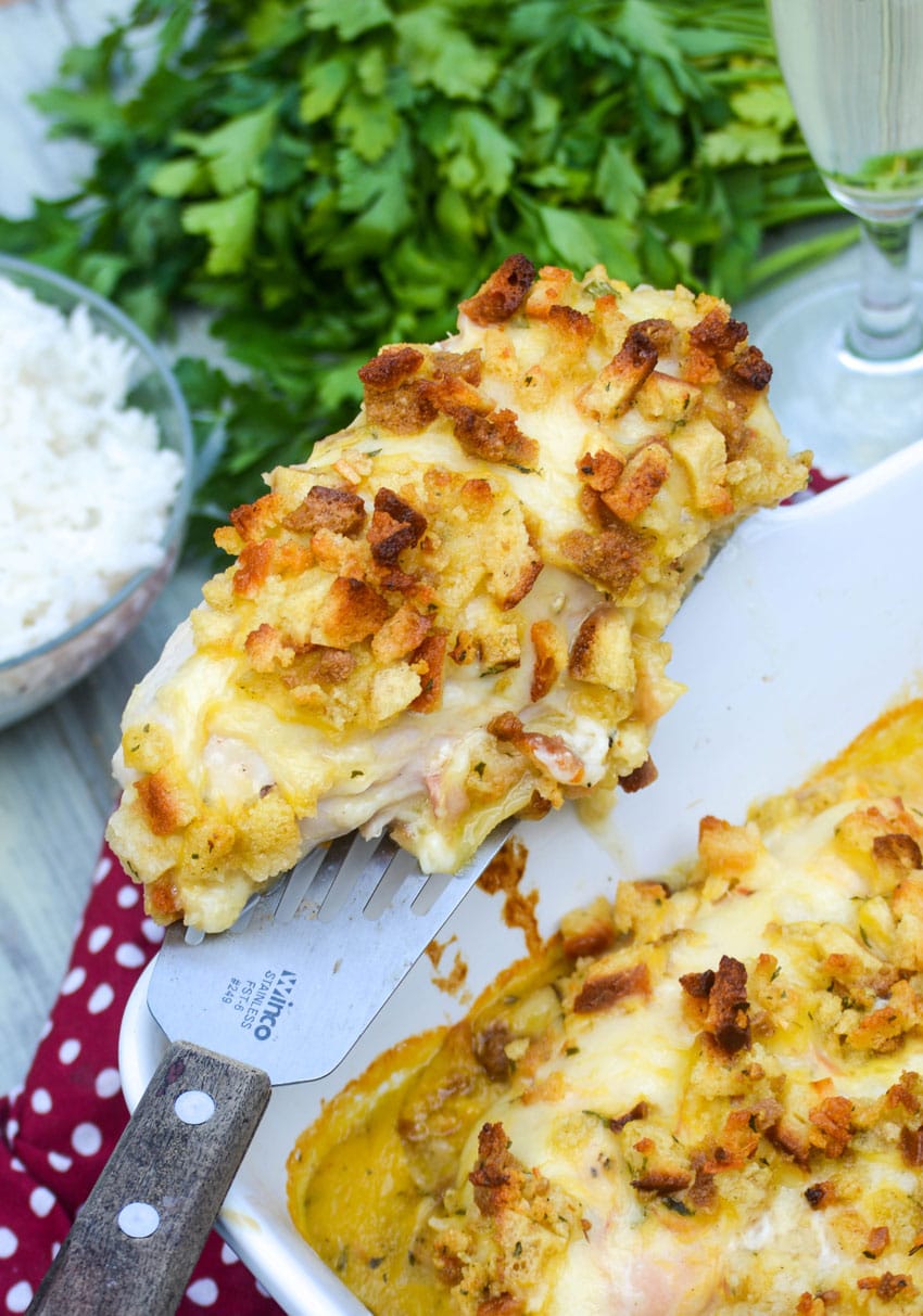 Easy Chicken In Wine Sauce Casserole a spatula holding a piece of herb crusted chicken in wine sauce & resting on the edge of a white casserole dish