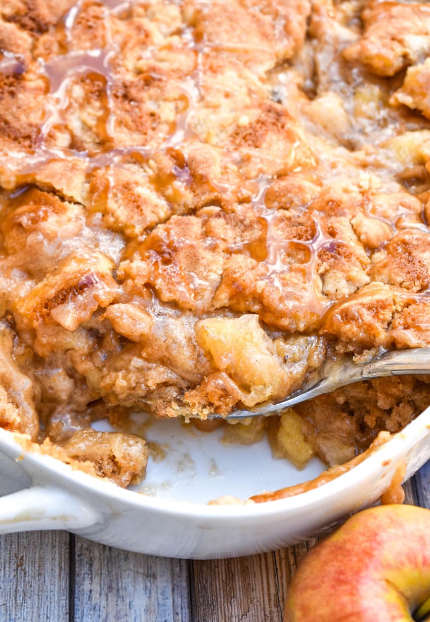 a silver spoon digging into a white baking dish filled with snicker doodle apple cobbler