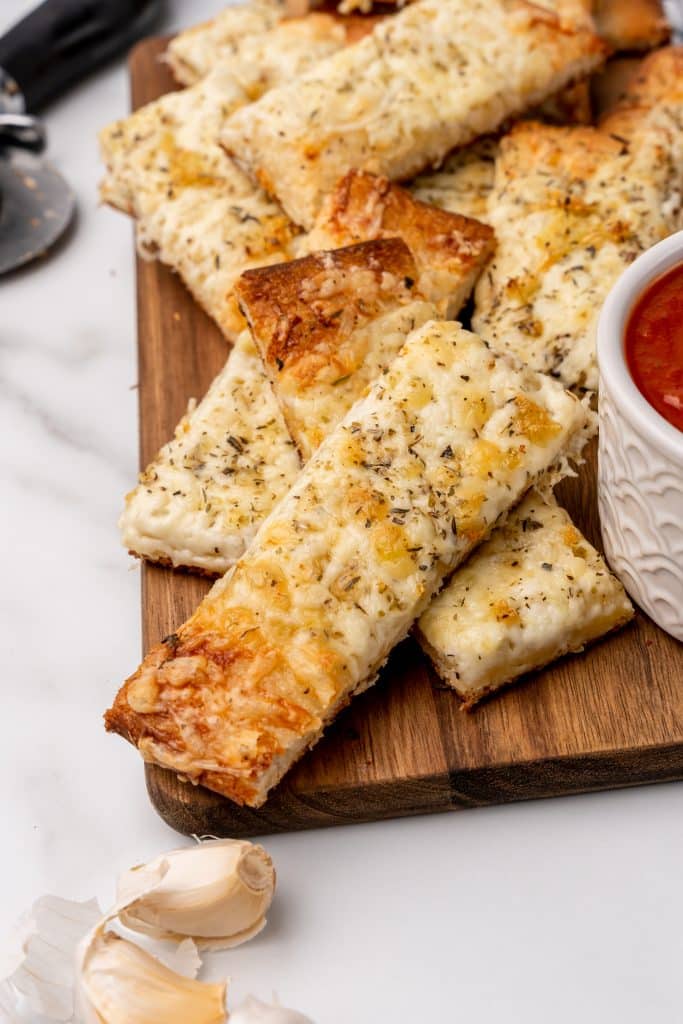 a pile of cheesy garlic breadsticks on a wooden cutting board next to a small bowl of marinara sauce
