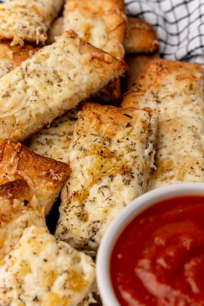 cheesy garlic breadsticks piled next to a small white bowl filled with marinara sauce