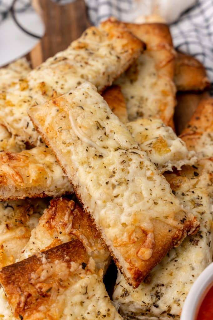 a pile of cheesy garlic breadsticks on a wooden cutting board next to a small bowl of marinara sauce