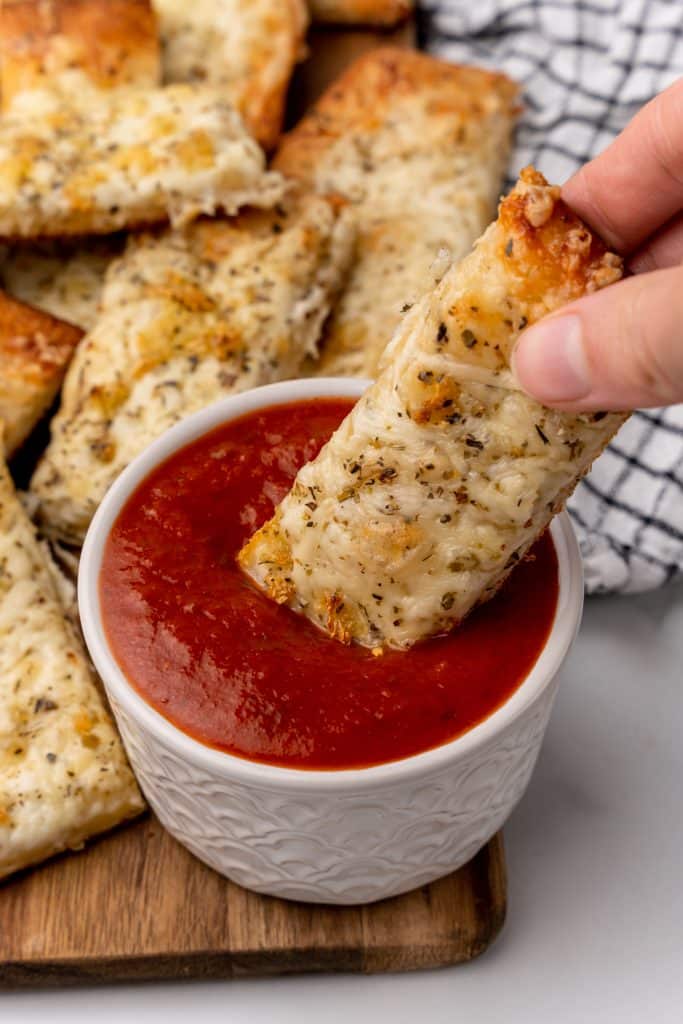 a hand dunking a cheesy garlic breadstick into a small white bowl filled with marinara sauce