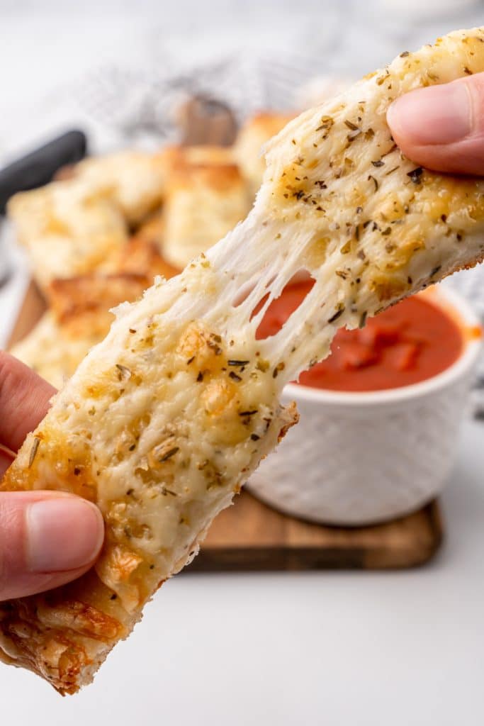 a hand puling a cheesy garlic breadstick in half to show stretching strings of melty cheese