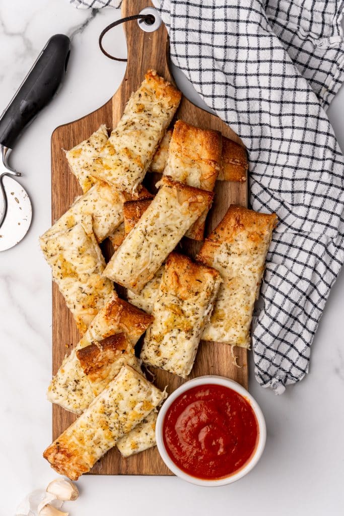 a pile of cheesy garlic breadsticks on a wooden cutting board next to a small bowl of marinara sauce