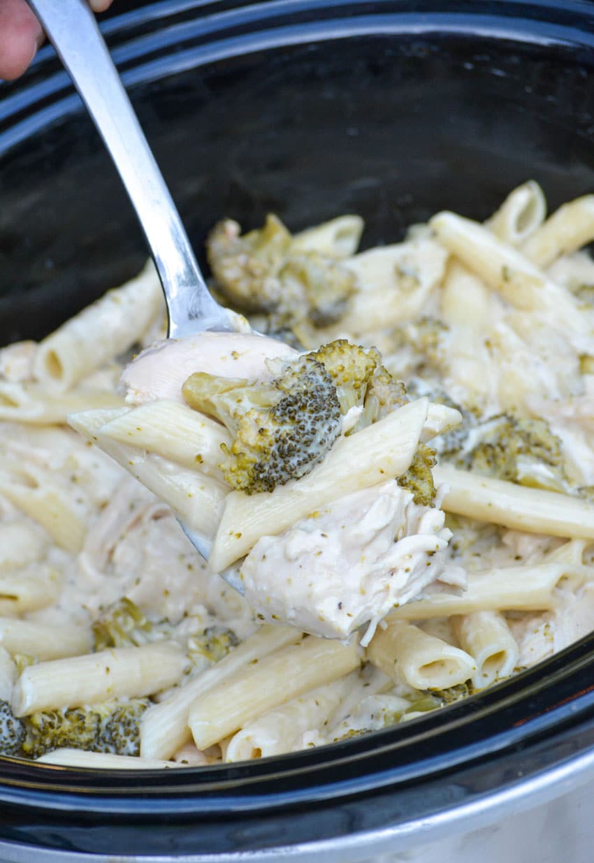a silver serving spoon holding a scoop of chicken alfredo over a slow cooker