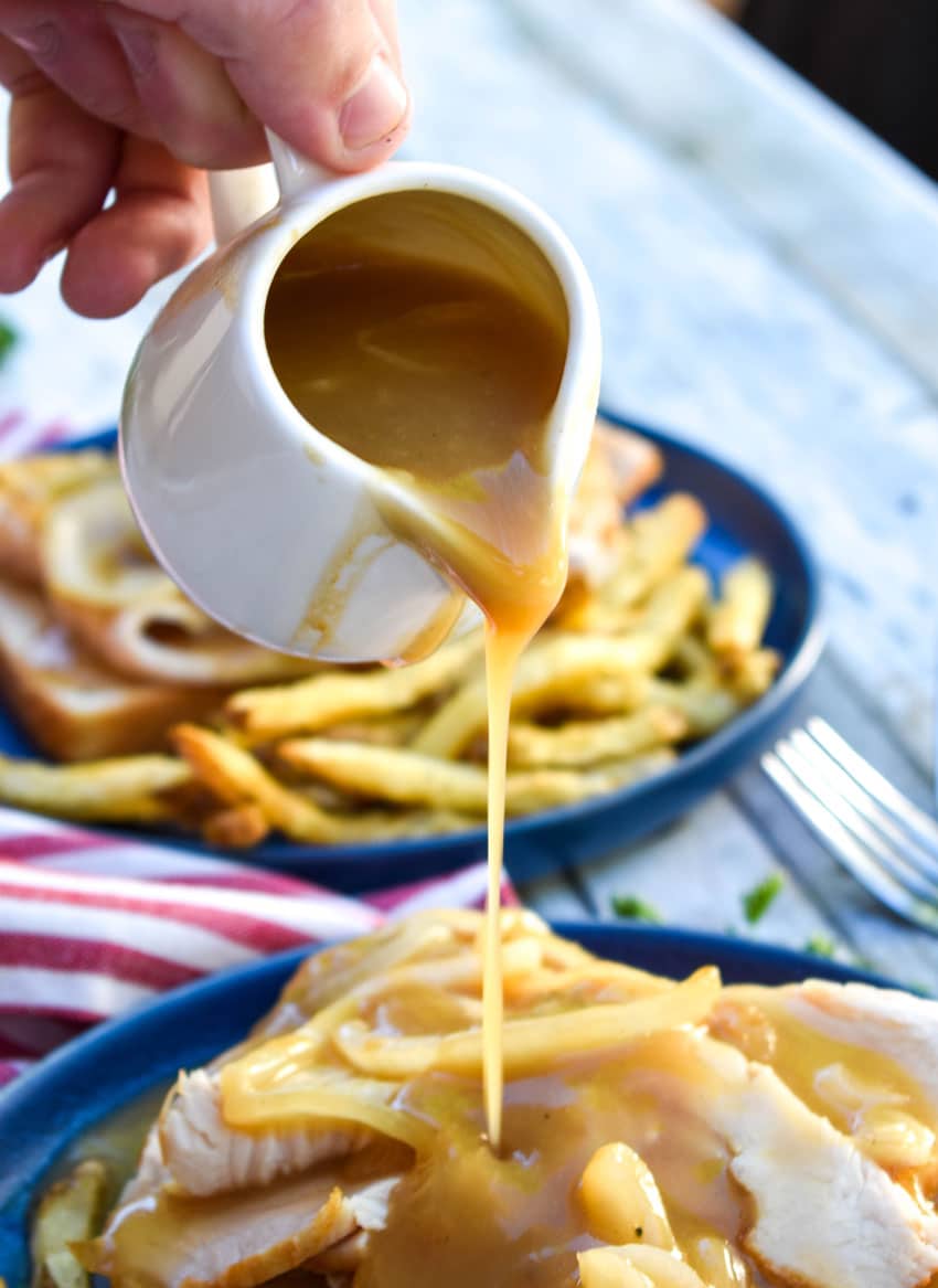 gravy being poured from a small white jar over an open faced turkey sandwich