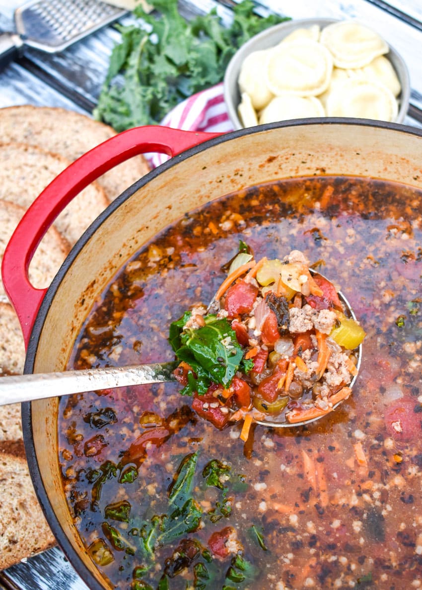 a silver ladle holding a scoop of sausage and lentil soup above a red Dutch oven