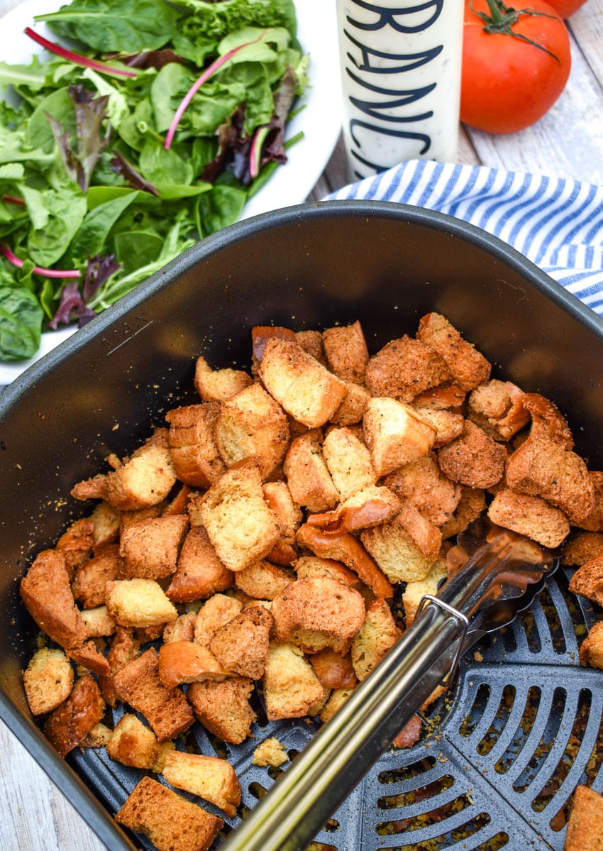 seasoned croutons made in the air fryer in the basket with a pair of silver tongs