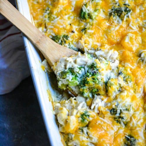 a wooden spoon digging into a cheesy chicken and rice casserole with broccoli in a white baking dish
