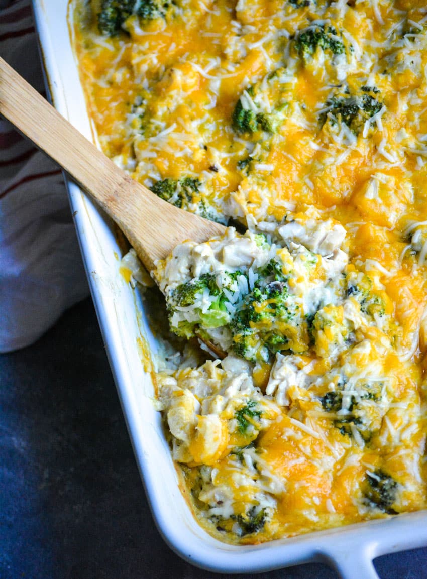 a wooden spoon digging into a cheesy chicken and rice casserole with broccoli in a white baking dish