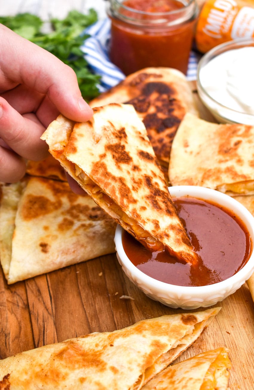 a hand holding up a cheesy chicken quesadilla above a small white bowl of mild taco sauce