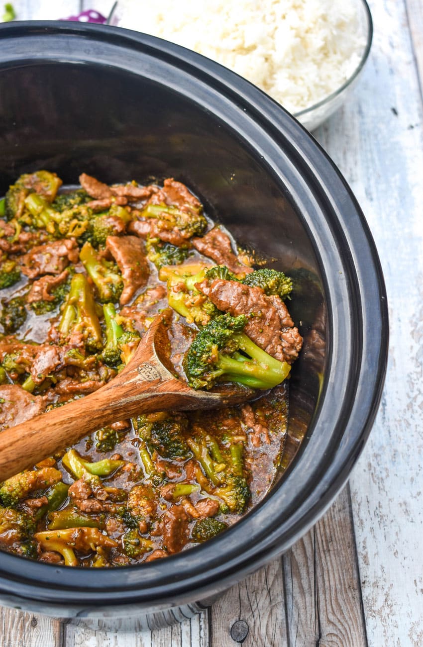 a wooden spoon holding a scoop of beef and broccoli in a black slow cooker
