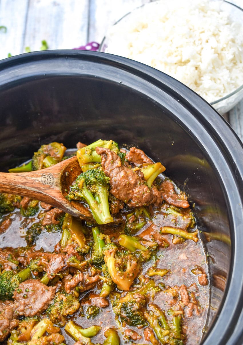 a wooden spoon holding a scoop of beef and broccoli in a black slow cooker