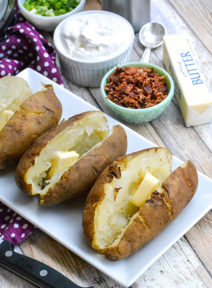 three buttered instant pot baked potatoes arranged in a row on a white serving platter