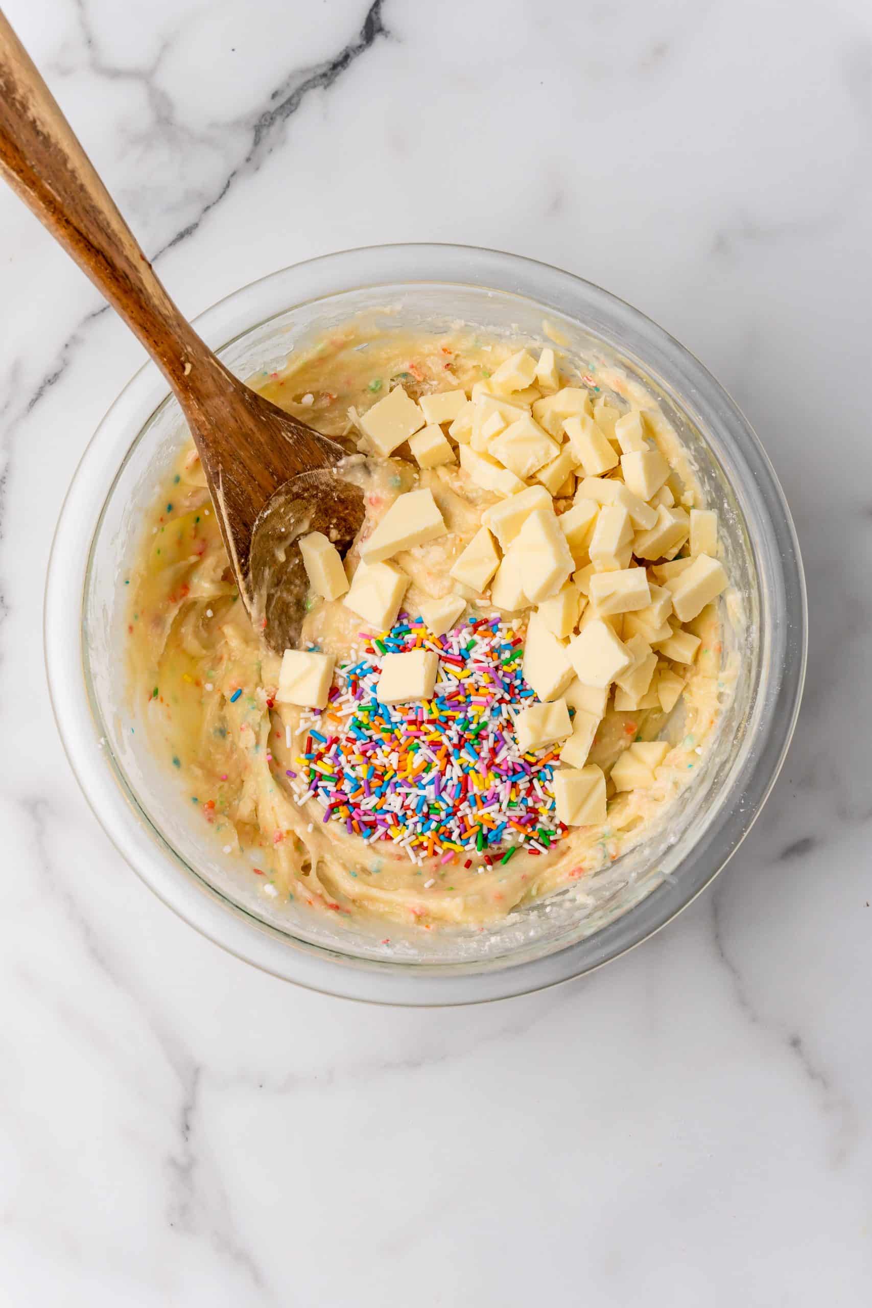 chunks of white chocolate and sprinkles in a glass bowl filled with cake batter