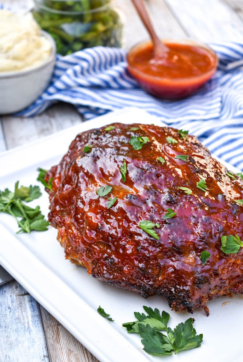a sauce covered air fryer meatloaf on a small white platter with fresh herbs in the corners