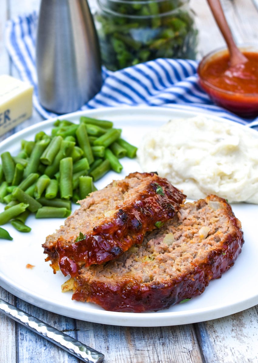 two slices of copycat cracker barrel meatloaf on a white dinner plate with green beans and mashed potatoes on the side