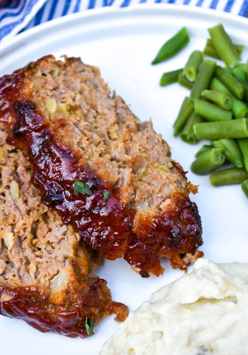 two slices of copycat cracker barrel meatloaf on a white dinner plate with green beans and mashed potatoes on the side