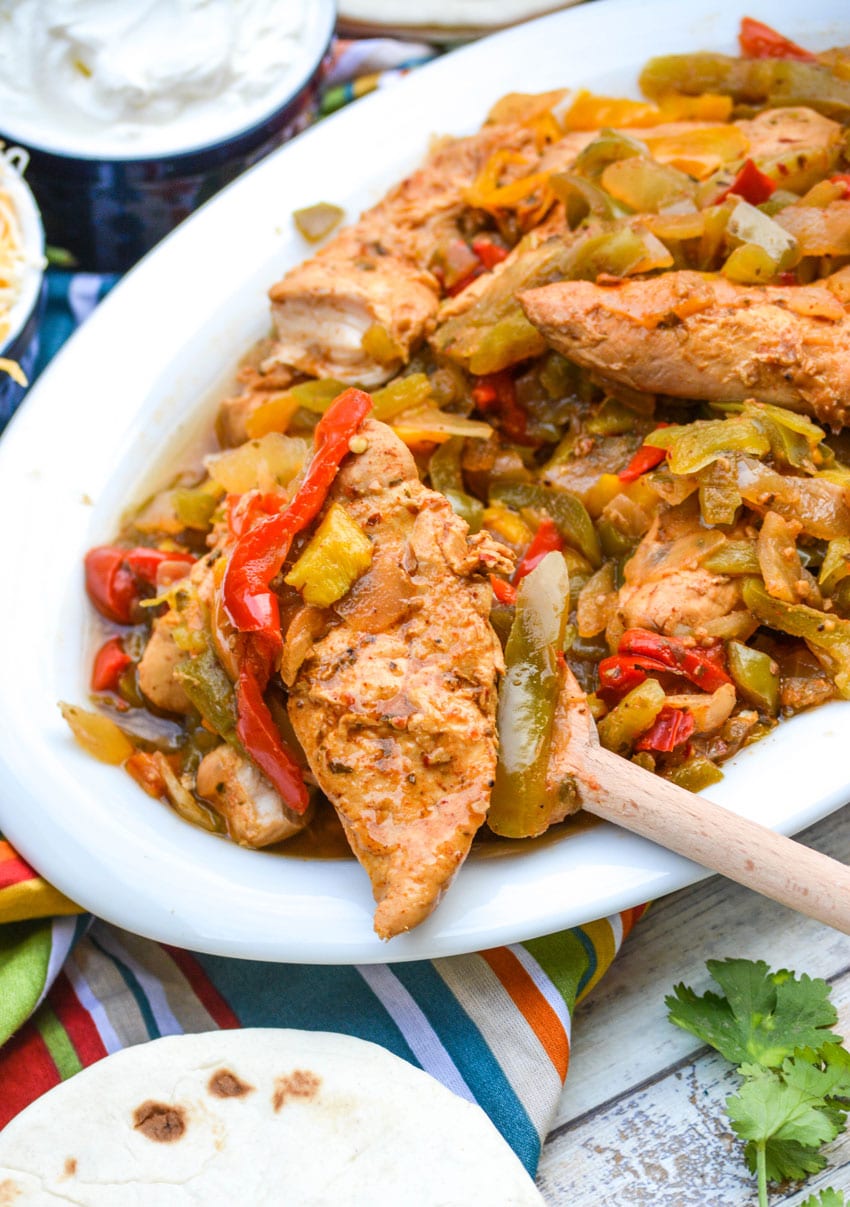 a wooden spoon scooping crockpot chicken fajitas out of a white serving bowl