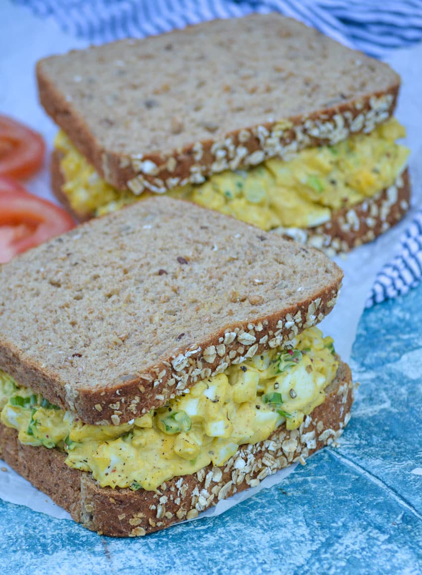 two sandwiches of curried egg salad on fluffy wheat bread sitting on a piece of white parchment paper