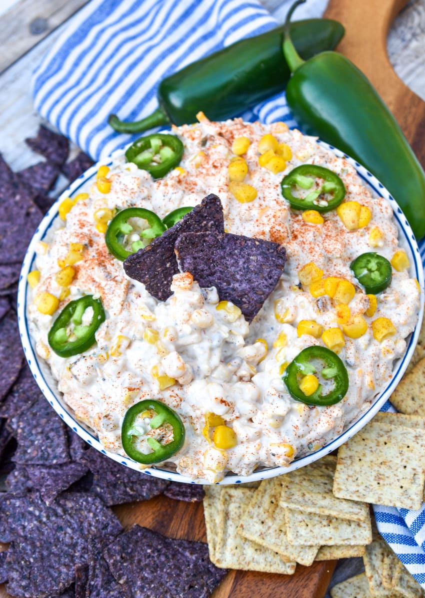 blue tortilla chips in a bowl of creamy corn dip topped with sliced jalapenos