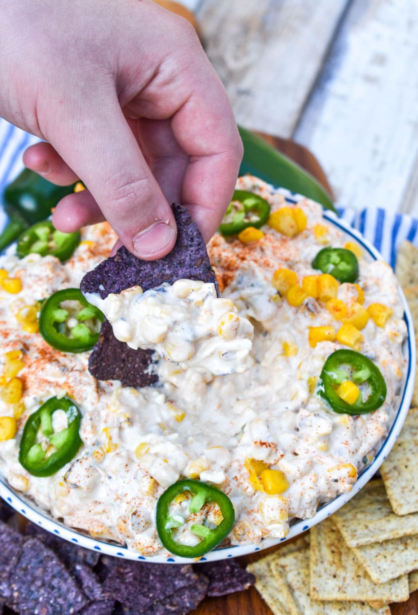 a hand holding up a blue corn chip topped with elote corn dip