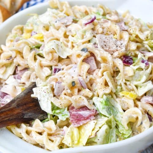 a wooden spoon stuck in a large white bowl filled with Italian Grinder Pasta Salad
