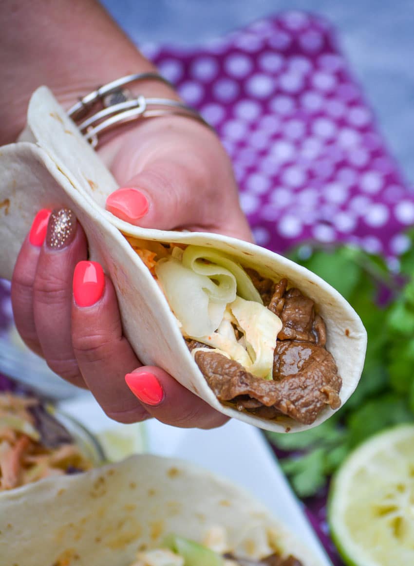 Korean Steak Tacos a hand holding up a korean stuffed taco in a flour tortilla