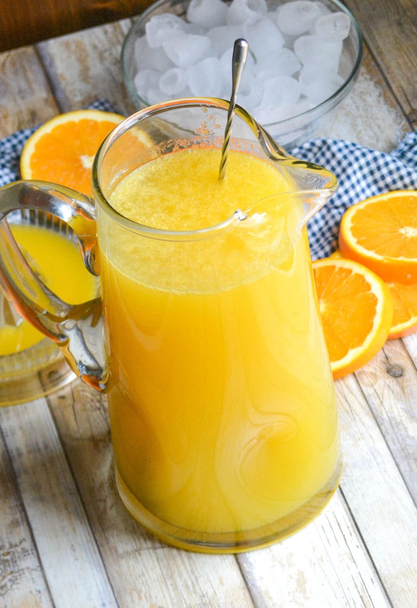 a homemade orangeade recipe in a large glass pitcher with a silver stirrer stuck in the middle
