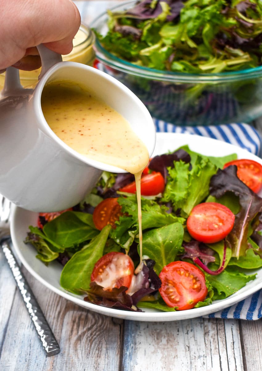a small white jar pouring honey mustard vinaigrette onto a simple green salad