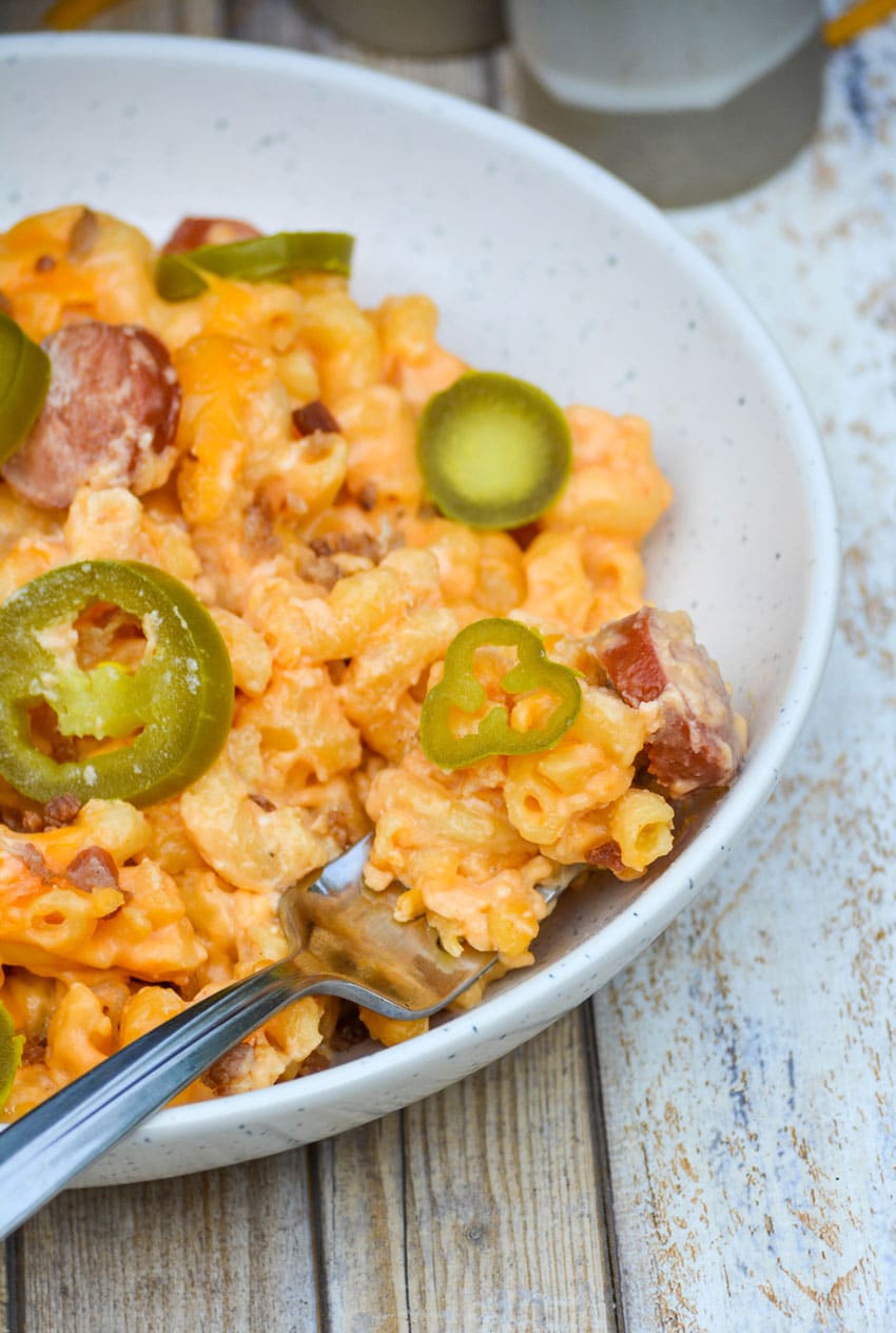 a silver fork stuck in a bowl of baked macaroni and cheese with hot dogs topped with crumbled bacon and slices of jalapeno