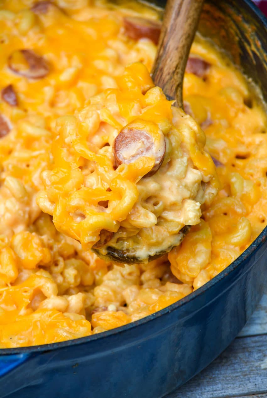 a wooden spoon scooping firehouse mac and cheese with hot dogs out of a large blue pot