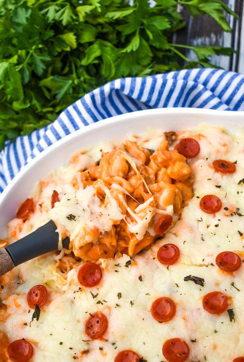a serving spoon scooping pizza mac and cheese out of a large white serving bowl