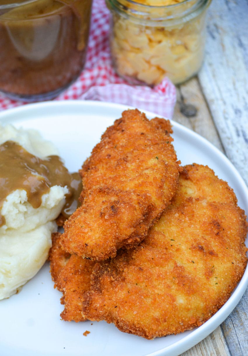 three pieces of potato flake chicken stacked on a white dinner plate with mashed potatoes and gravy on the side