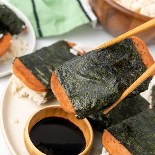 wooden chop sticks holding a piece of spam misubi above a wooden dipping bowl filled with Asian sauce