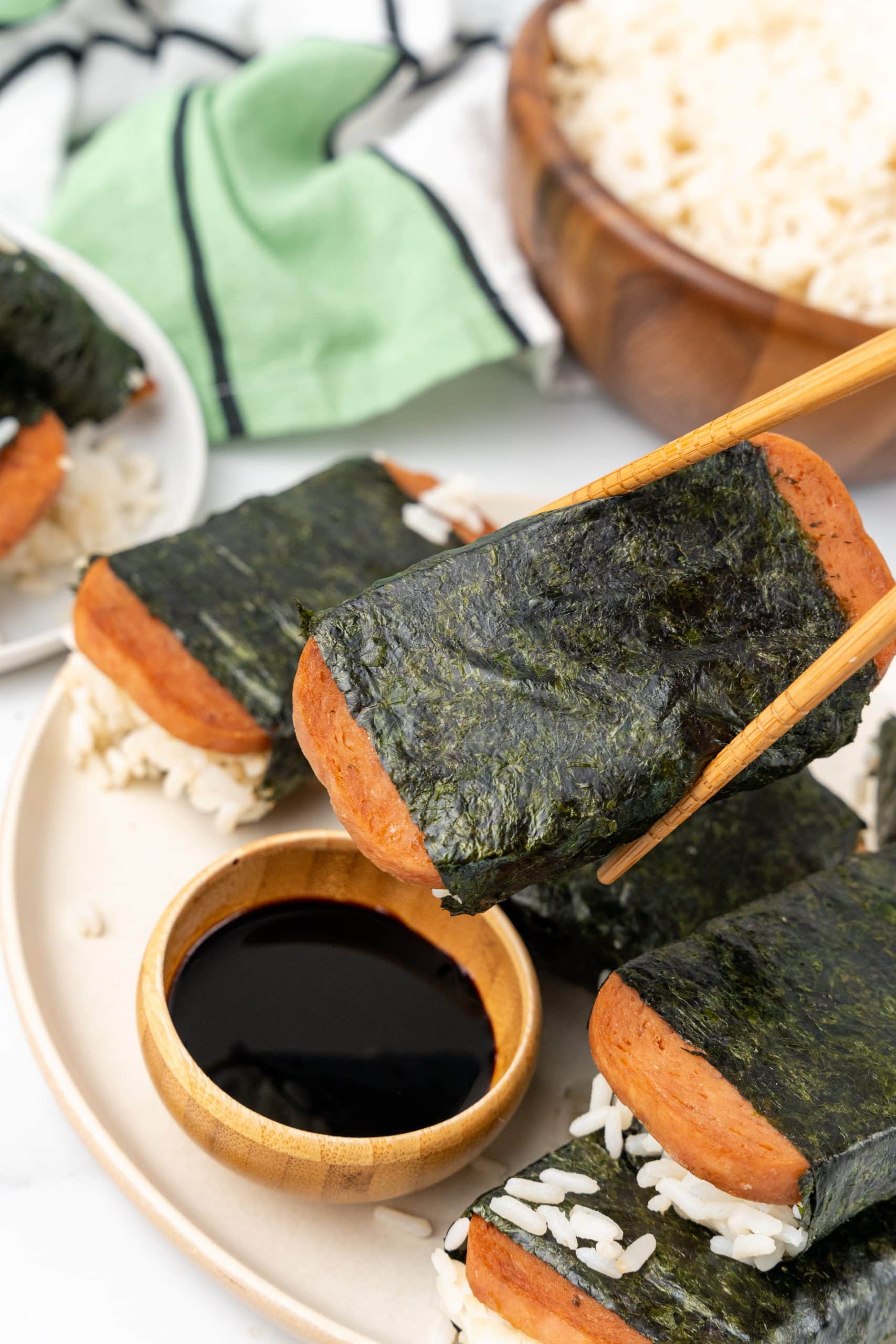 wooden chop sticks holding a piece of spam misubi above a wooden dipping bowl filled with Asian sauce