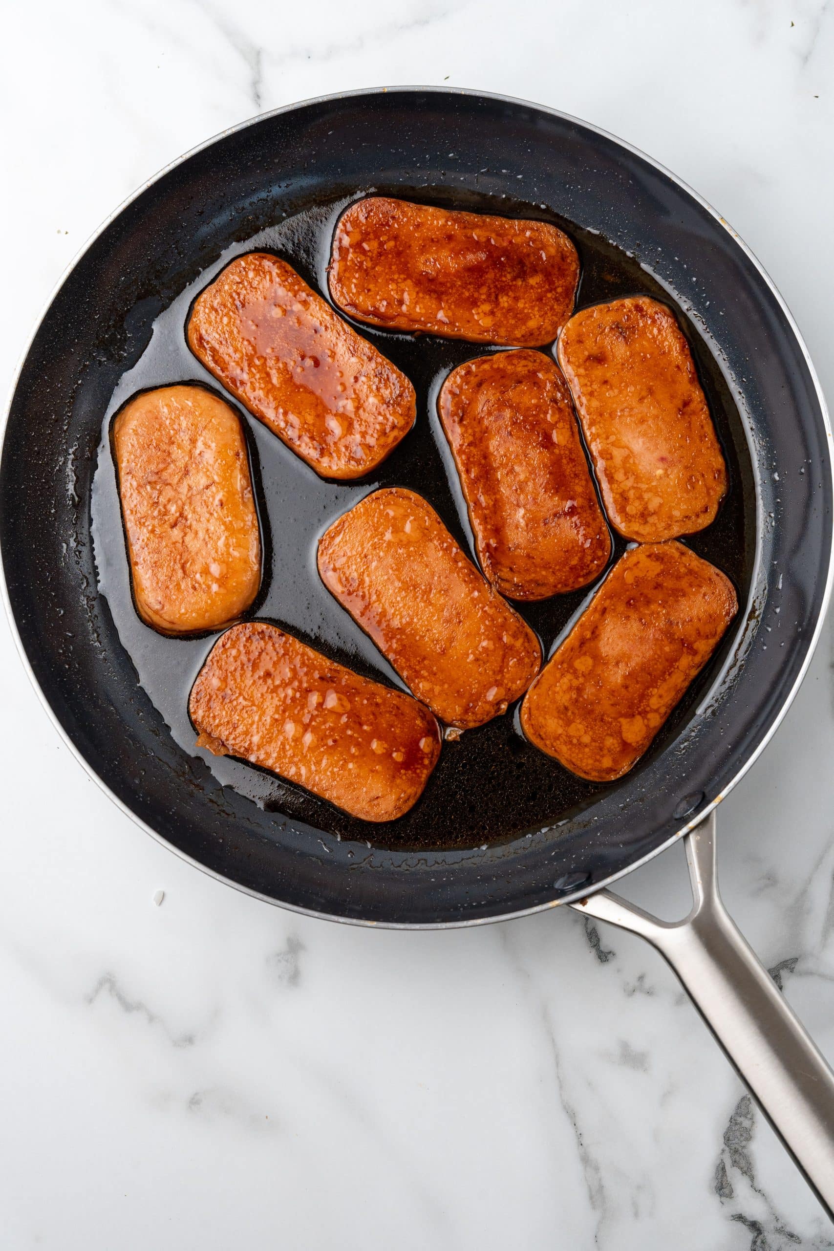 cooked slices of spam coated in sauce in a large black skillet