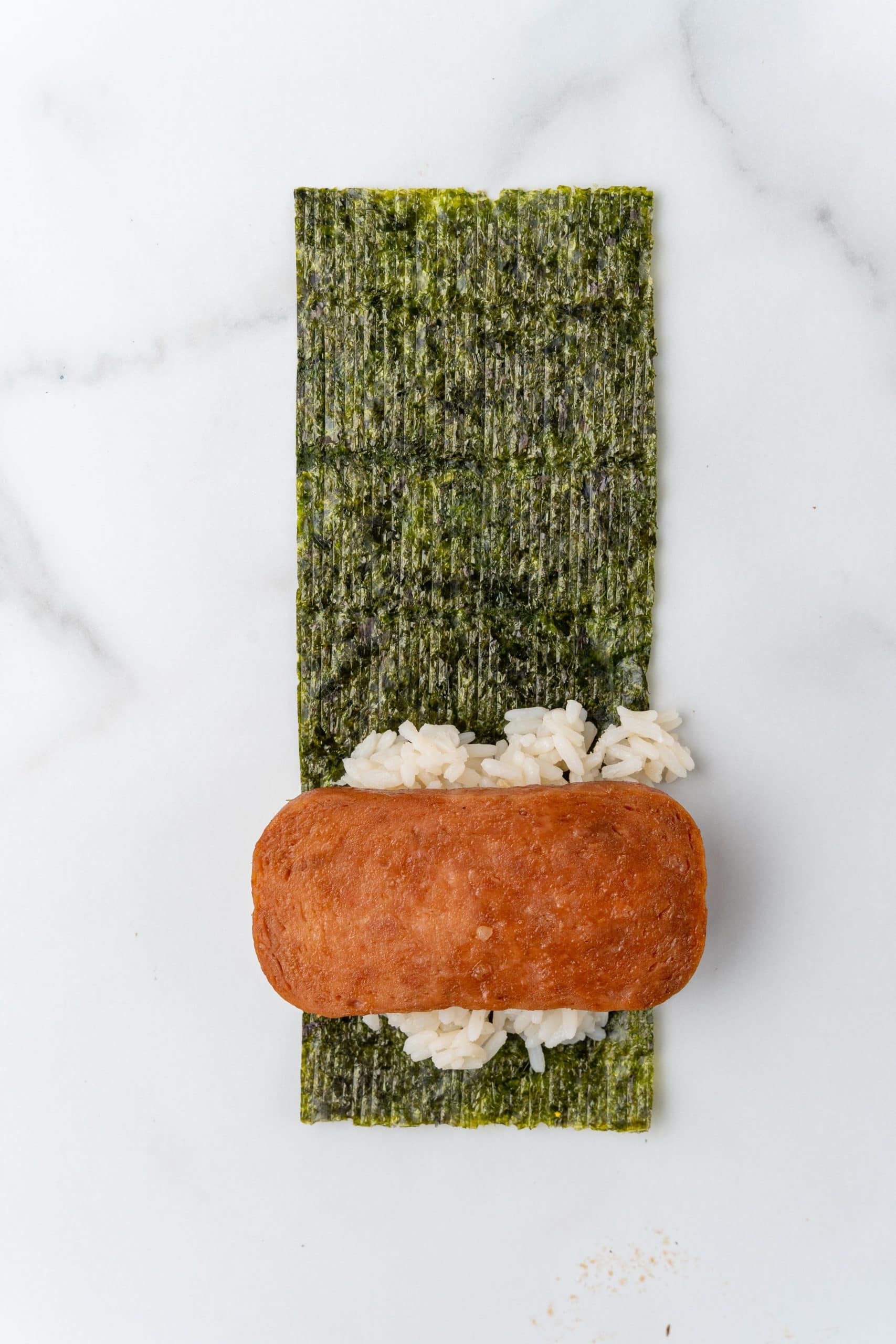a slice of spam on top of steamed white rice on the edge of a sheet of nori