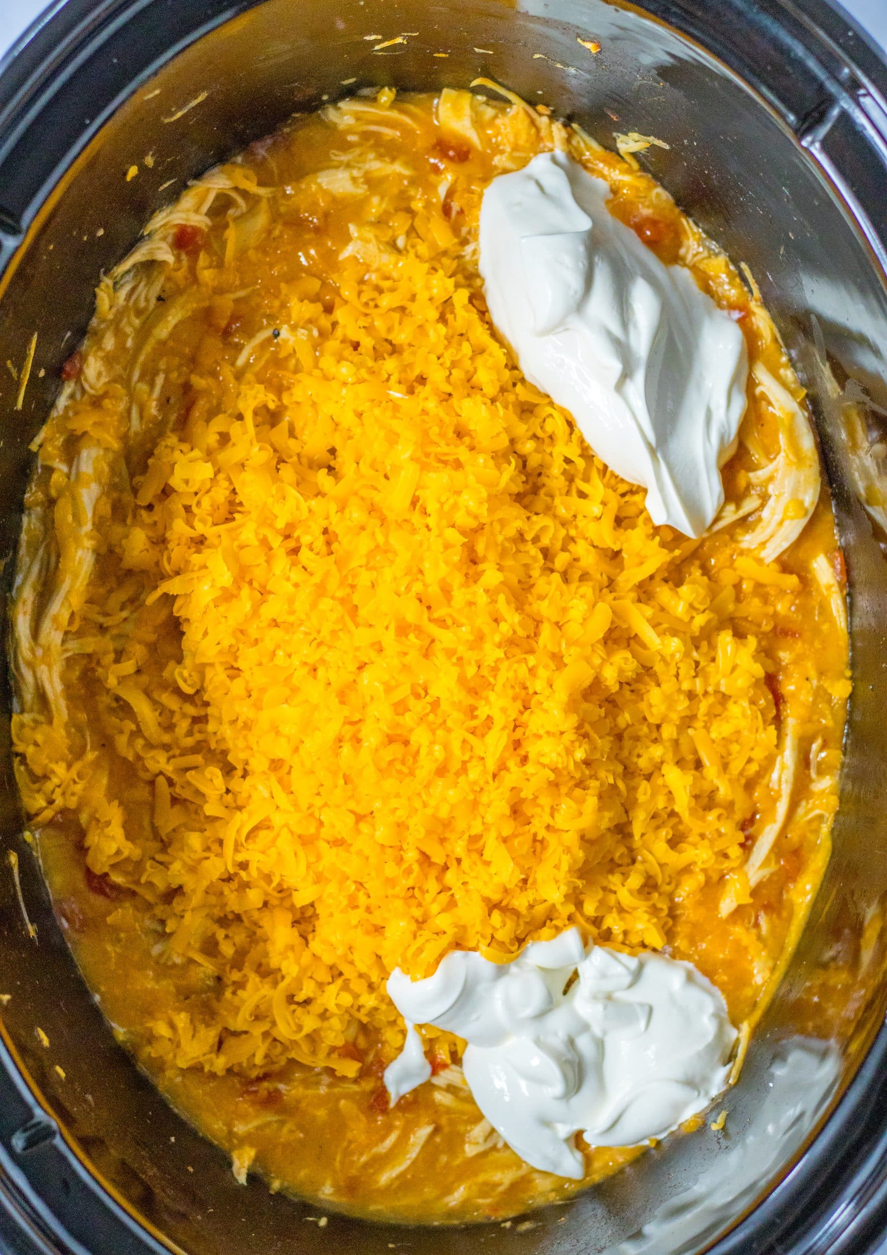 shredded cheddar and sour cream being added to cheesy chicken in the black bowl of a slow cooker