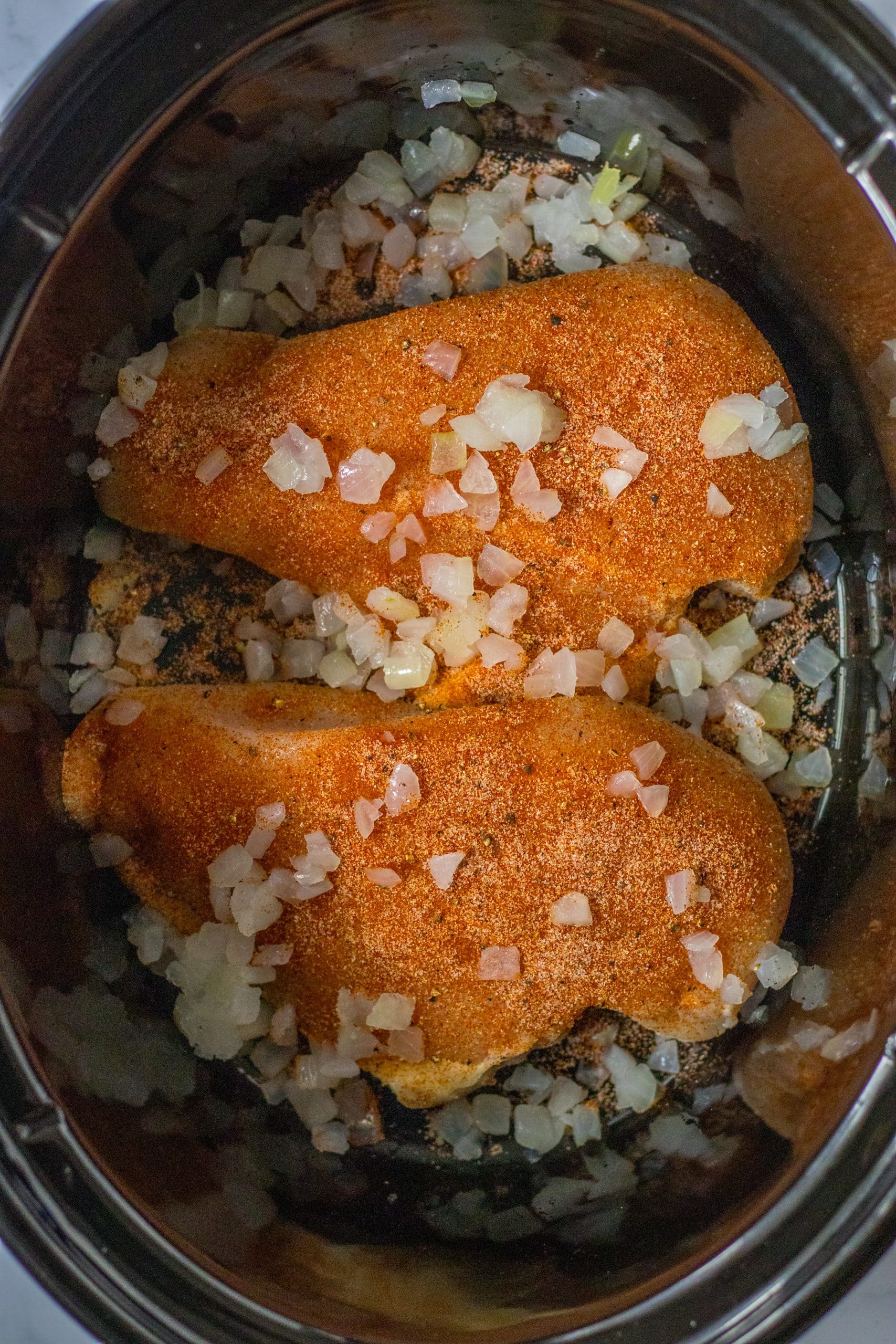 seasoned chicken breasts and diced onions in the black bowl of a slow cooker