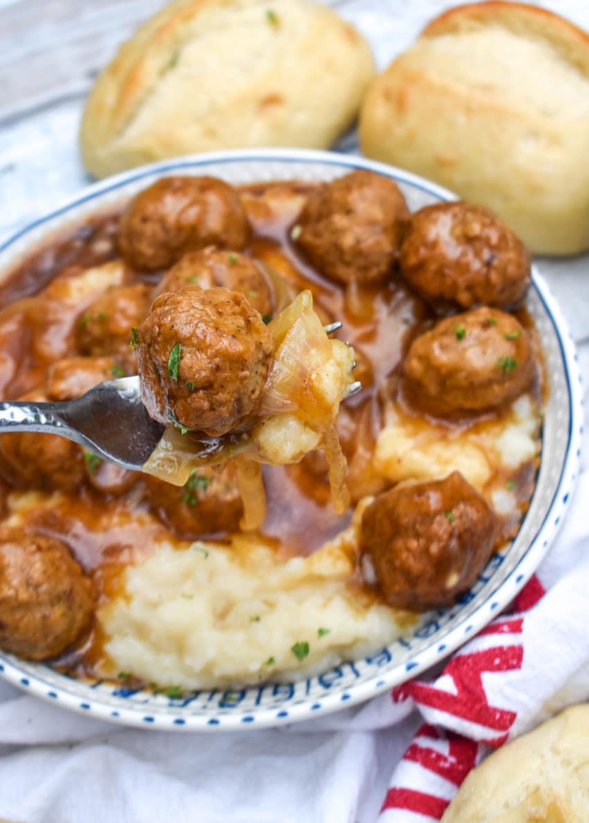 a silver fork holding up a scoop of salisbury steak meatballs in gravy with mashed potatoes over a blue and white bowl