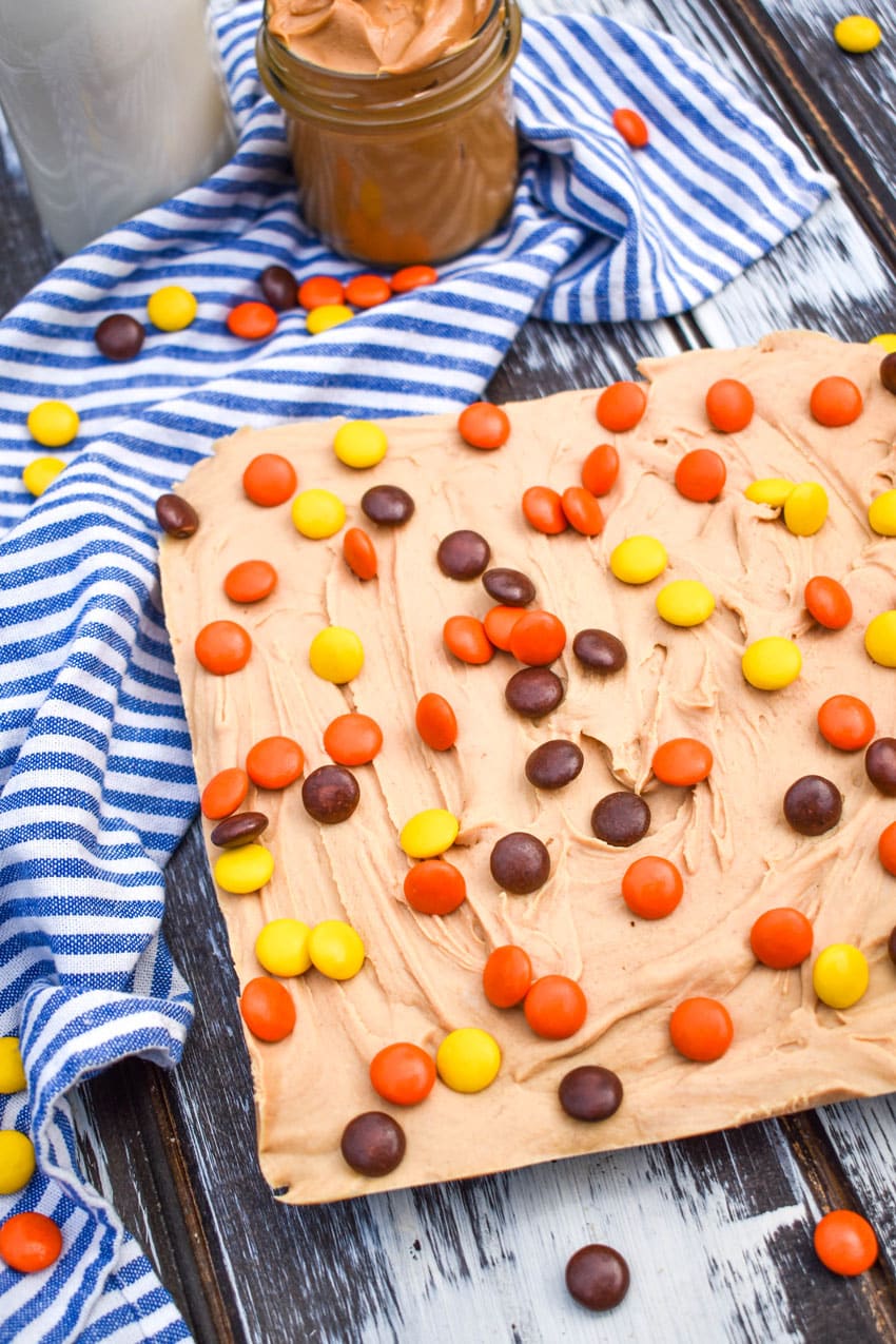 a block of easy peanut butter fudge on a wooden table surrounded by Reese's pieces candy