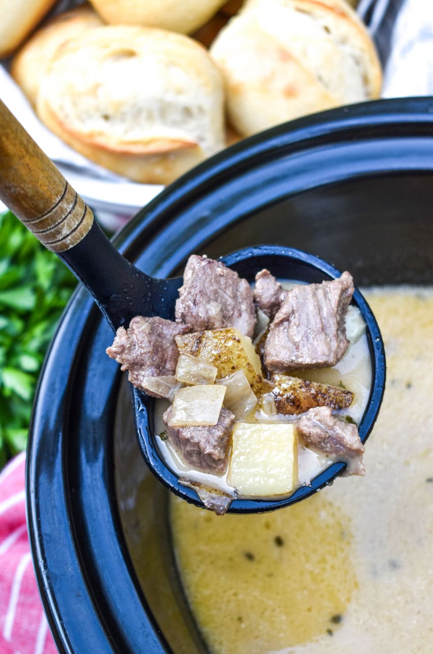 a ladle scooping creamy steak and potato soup out of a black crockpot