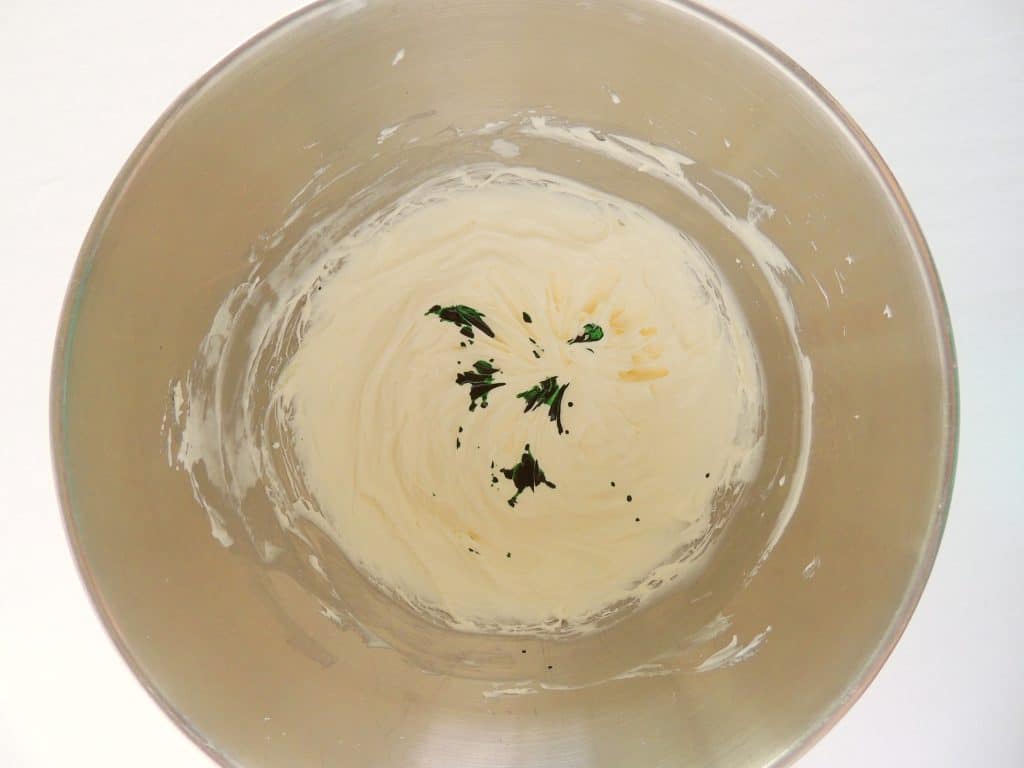 drops of green food coloring in marshmallow fluff in a metal mixing bowl