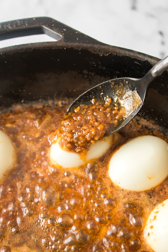 a silver spoon spooning sauce over a hard boiled egg in a black skillet