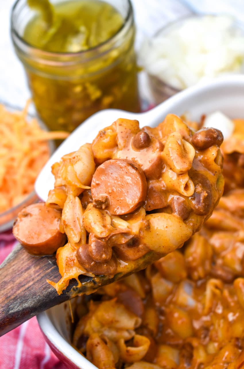 a wooden spoon holding up a scoop of chili dog mac and cheese with hot dogs