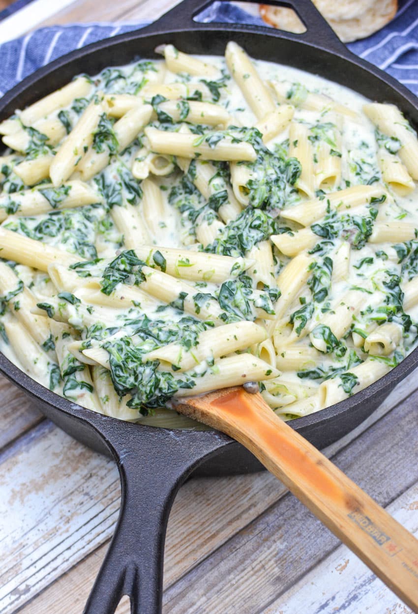 a wooden spoon stuck in a cast iron skillet filled with creamed spinach mac and cheese