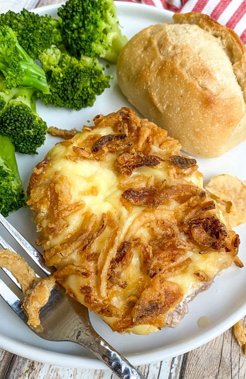 french onion pork chops on a white plate with steamed broccoli and a dinner roll on the side