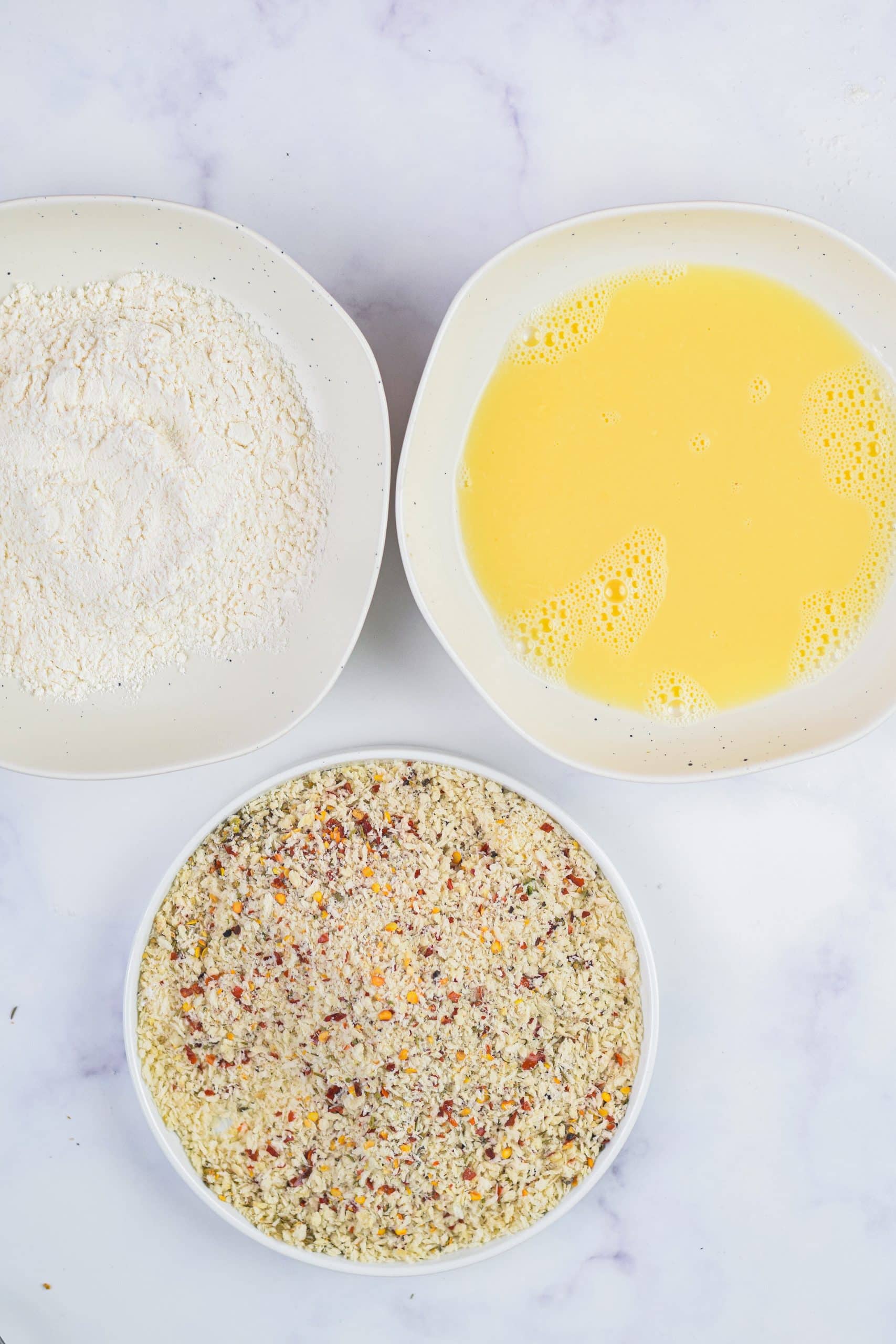 three white bowls, filled with flour, another seaasoned bread crumbs, and the last beaten eggs